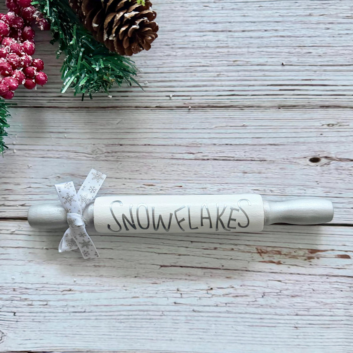A rustic wooden rolling pin labeled 'SNOWFLAKES' in black lettering, tied with a white patterned ribbon, placed on a whitewashed wooden surface with holiday greenery and red berries in the corner.