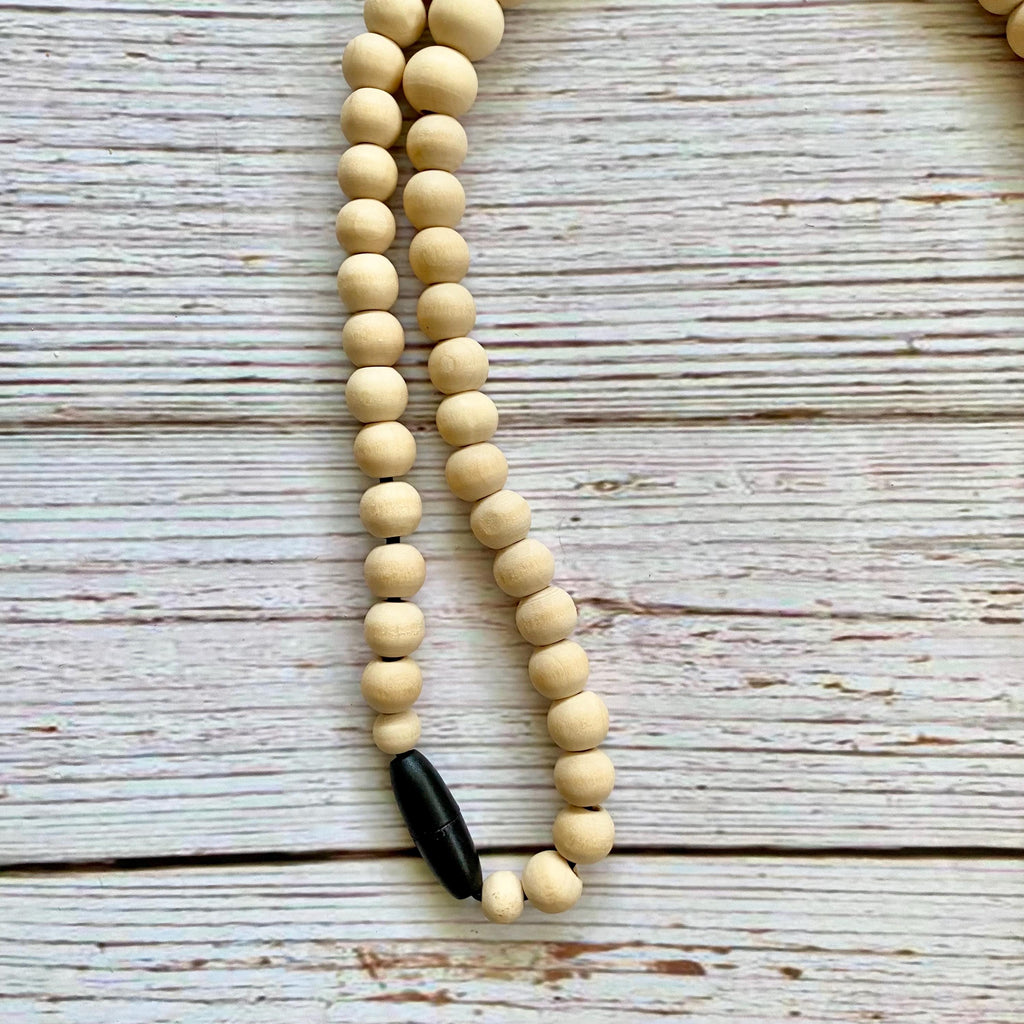 A beaded necklace made of light natural wood beads with a black breakaway clasp at the bottom, laid on a whitewashed wooden surface.