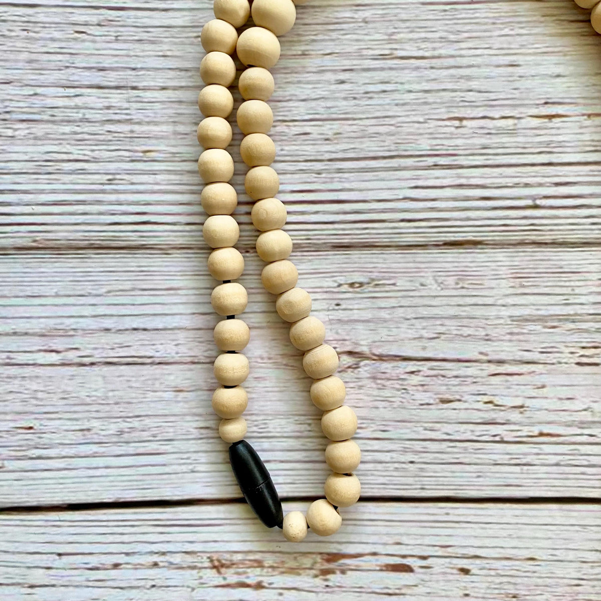 A beaded necklace made of light natural wood beads with a black breakaway clasp at the bottom, laid on a whitewashed wooden surface.