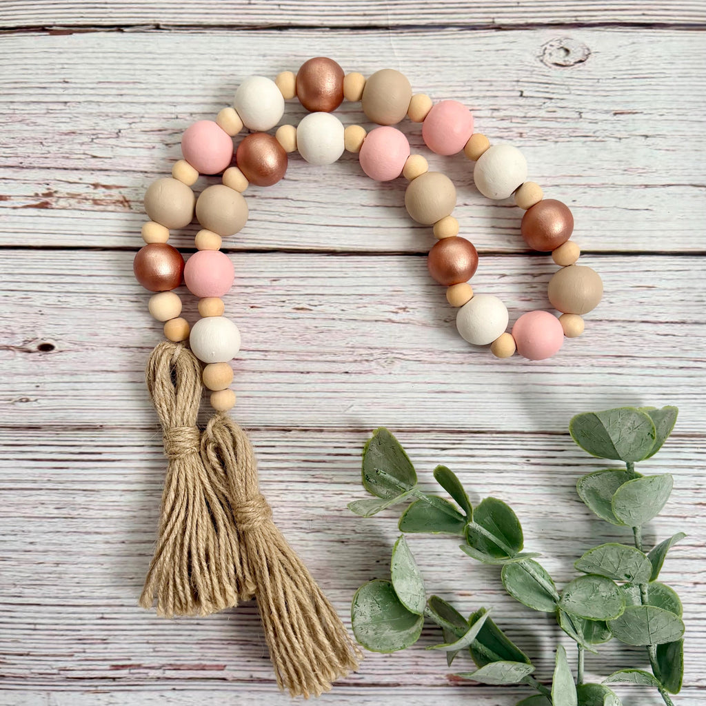 A beaded garland with alternating matte beige, white, and pink wooden beads, metallic rose-gold and copper beads, and two large tan jute tassels, arranged on a whitewashed wood surface with green eucalyptus leaves.