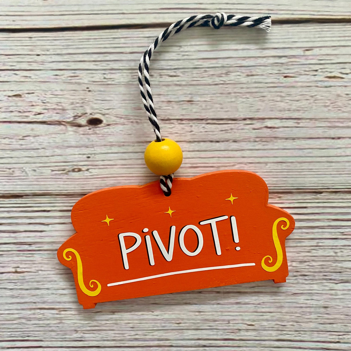 An orange wooden ornament shaped like a couch, with the word 'PIVOT!' in white letters and yellow decorative accents, hanging from a black-and-white twisted string with a yellow bead, set on a light wood plank background.