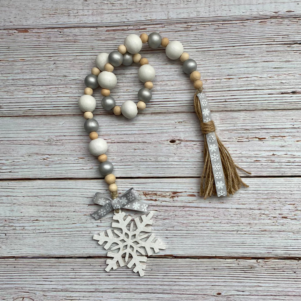 A beaded holiday garland with white, gray, and natural wood beads, a silver glitter tassel, a silver glitter bow, and a white wooden snowflake ornament, displayed on a rustic white-washed wooden background.