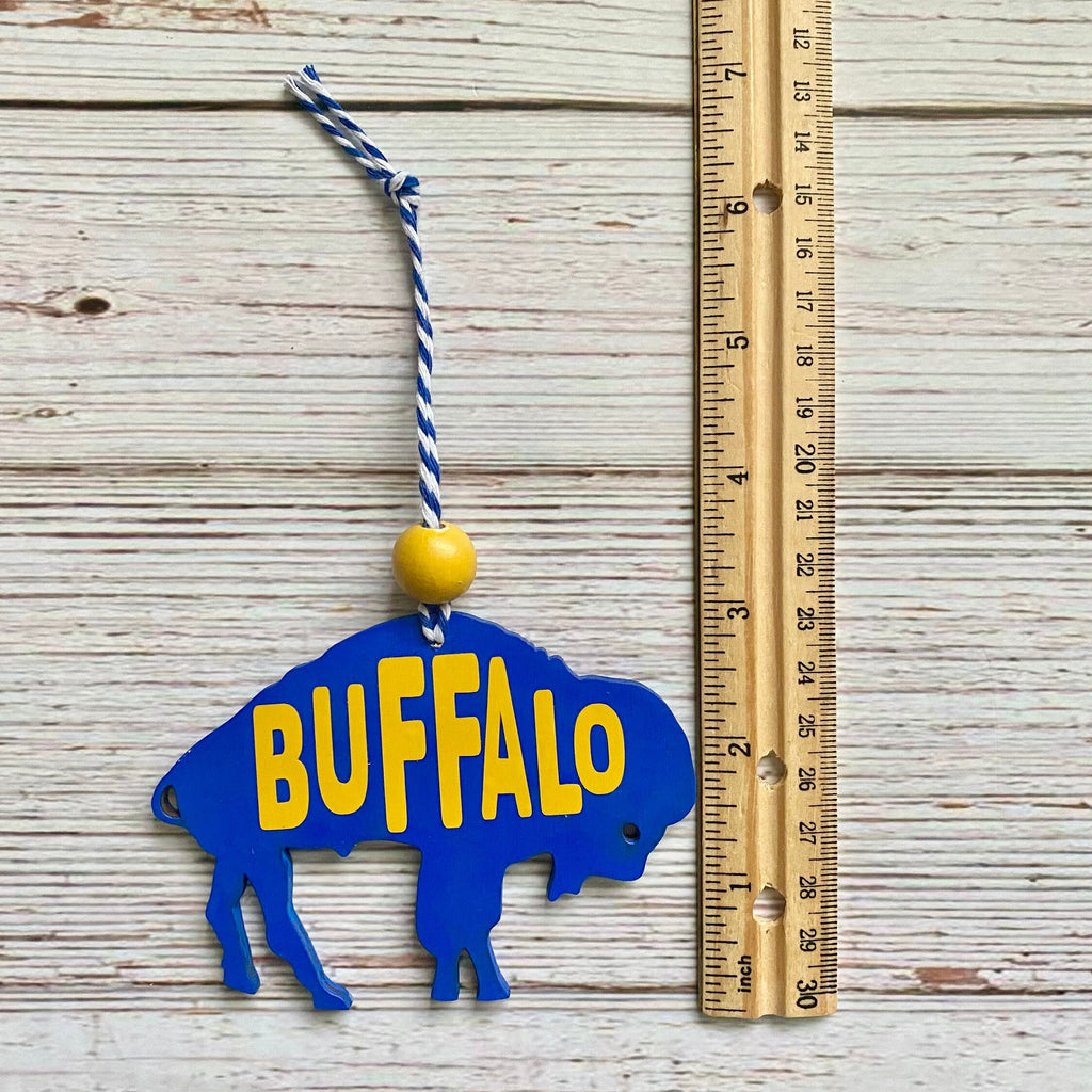 A blue wooden Buffalo ornament with yellow lettering, hanging by a blue and white string next to a wooden ruler on a whitewashed wood plank background.
