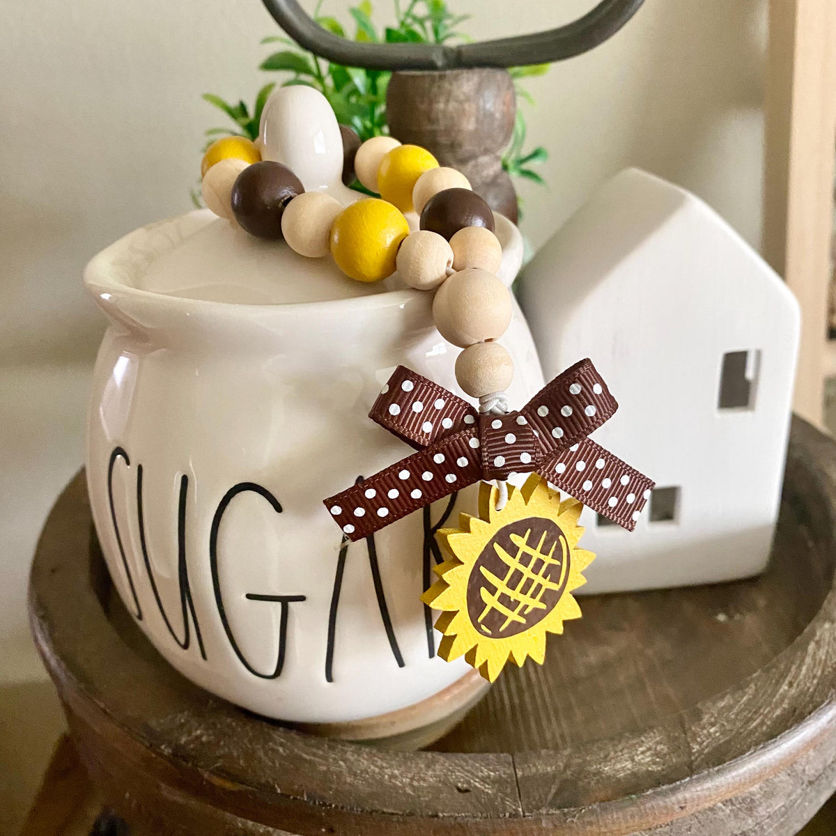 A white ceramic sugar canister with 'SUGAR' written on the front, styled with a wooden bead garland, a brown polka-dot ribbon bow, and a yellow sunflower ornament, sitting on a rustic wooden table next to a small white house figurine.