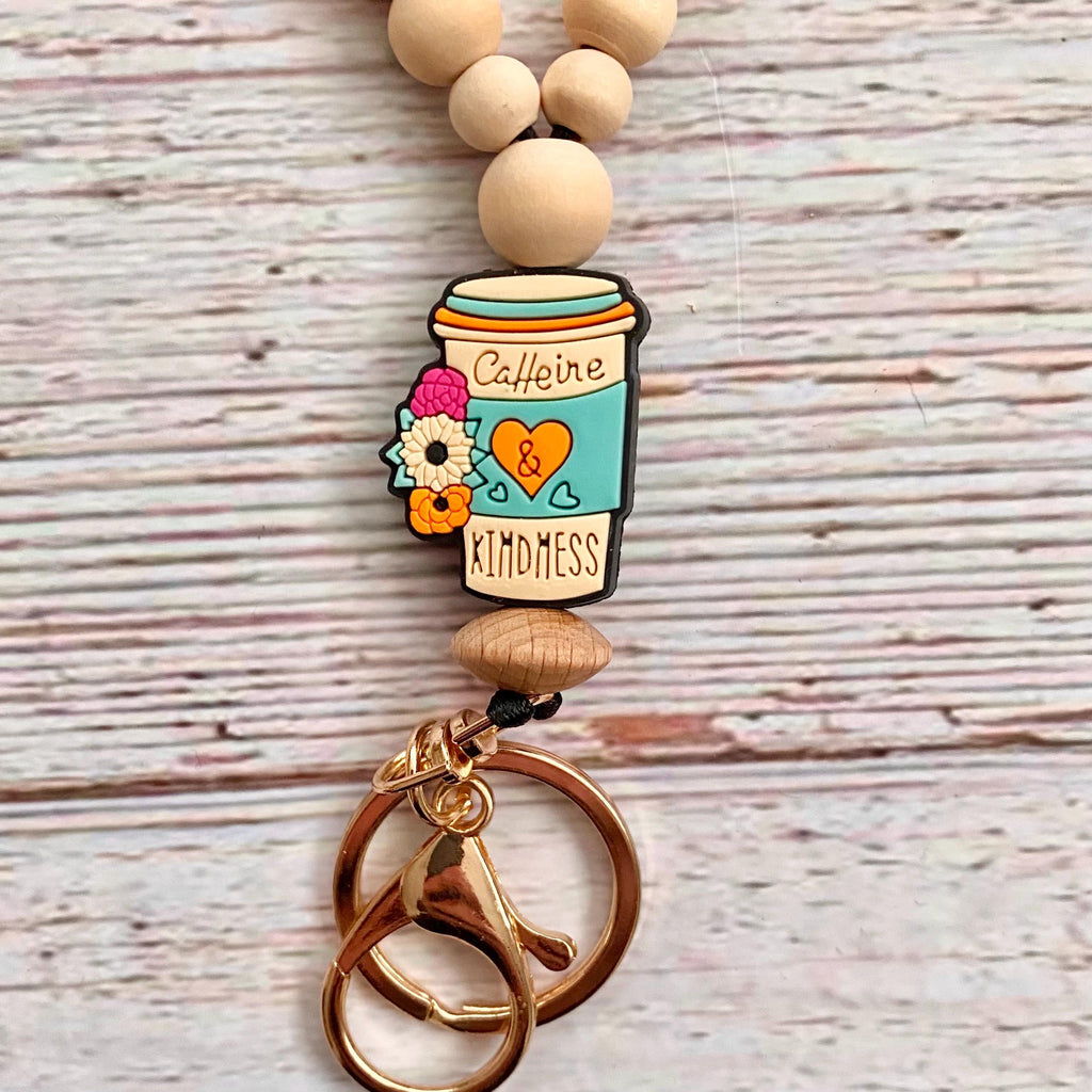 A beaded keychain with light wood beads, a coffee cup charm decorated with flowers and the words 'Caffeine' and 'KINDNESS', and two interlocking rose gold key rings on a whitewashed wood background.