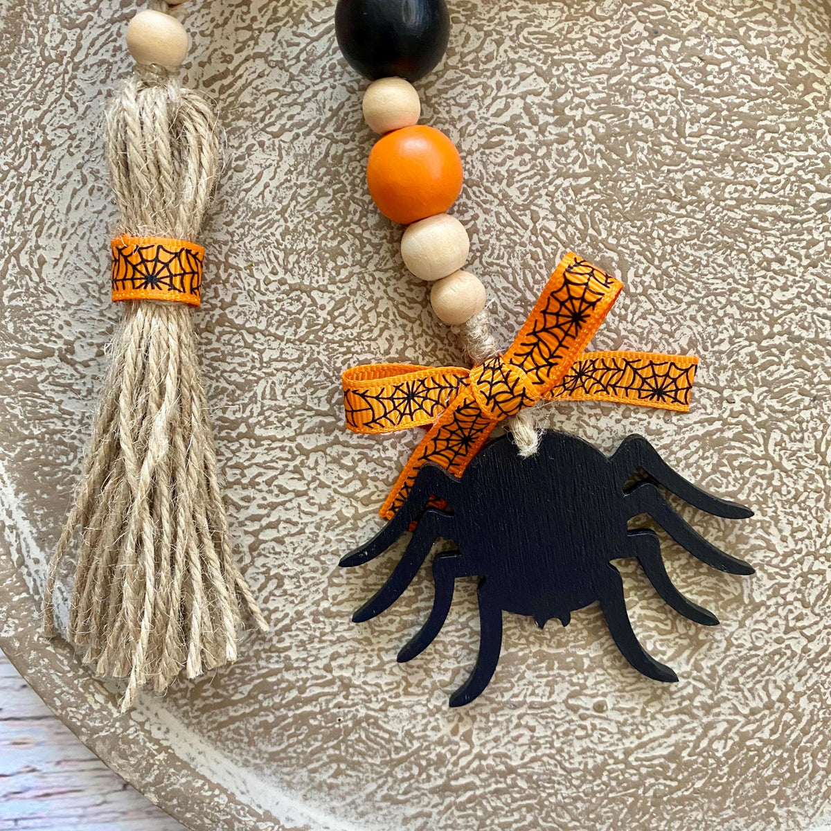 Halloween decorative garland with a black wooden spider cutout, orange patterned ribbon bow, white and orange beads, and a beige jute tassel on a textured beige surface.