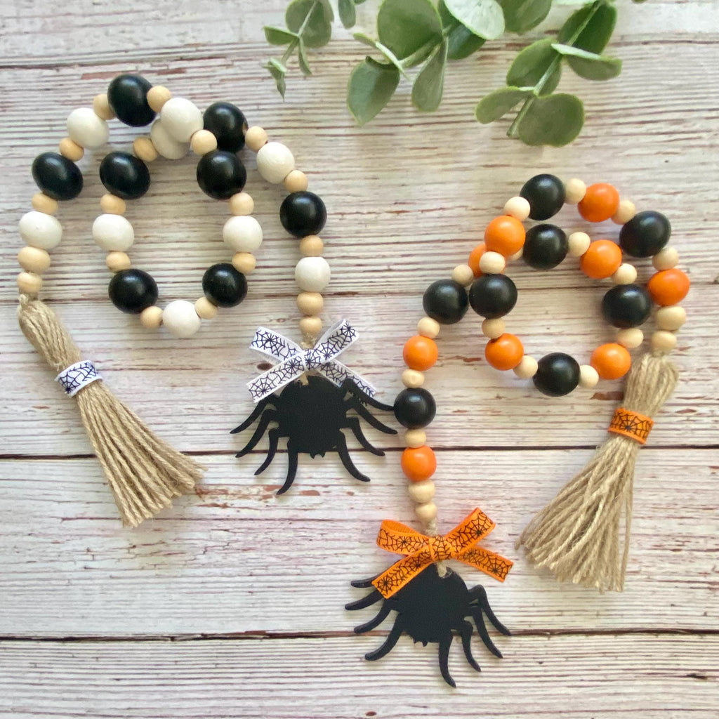 Two beaded Halloween garlands with black and natural wood beads, orange beads, jute tassels, and black spider cutouts, displayed on a whitewashed wooden surface with green leaves.