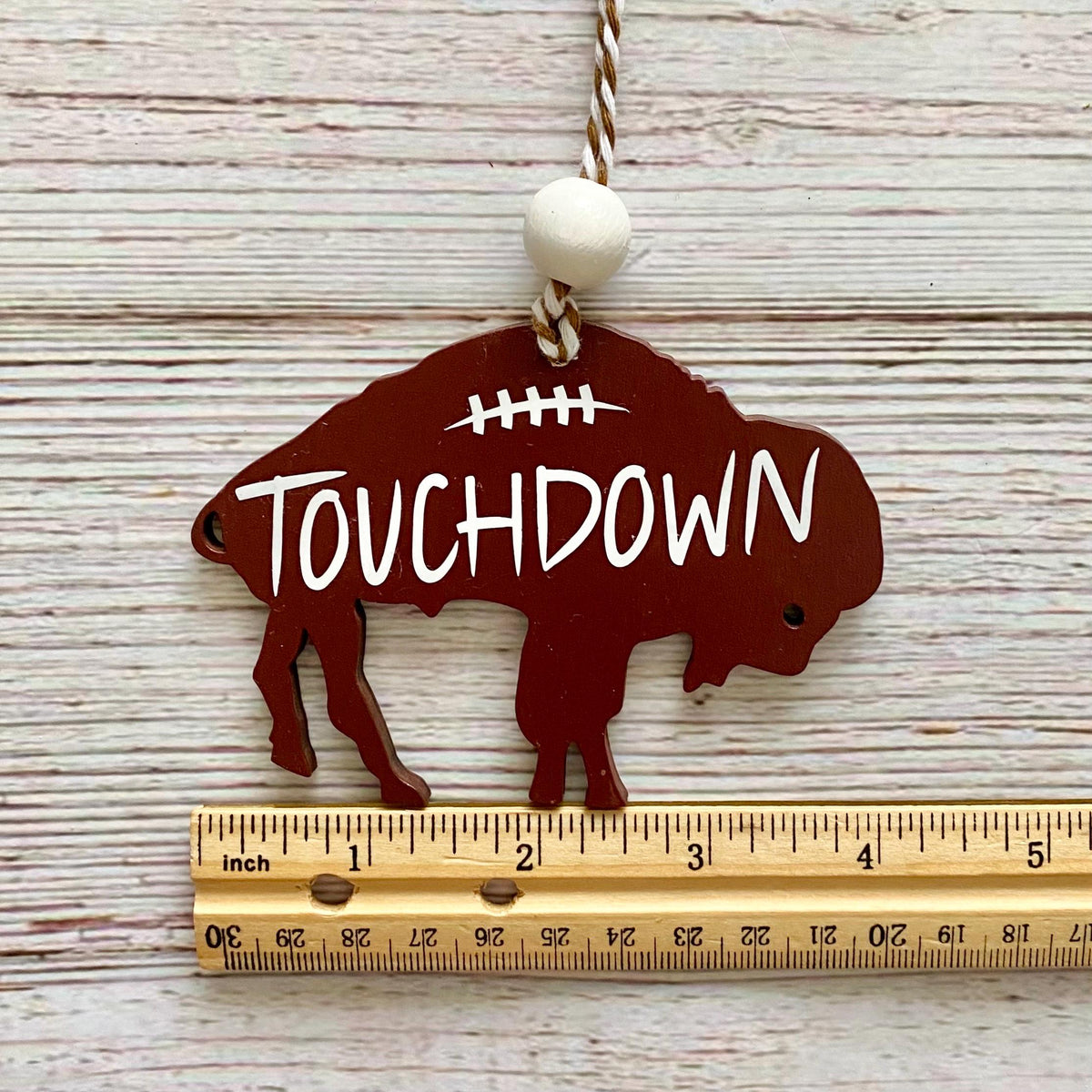 A buffalo-shaped metal ornament painted dark red with 'TOUCHDOWN' in white lettering, hanging from a rope with a white bead. A wooden ruler is placed below it to show scale on a whitewashed wood background.