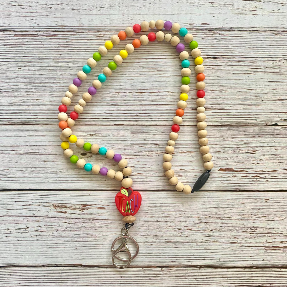 A beaded necklace with natural wood beads, colorful rainbow accents, and a red heart-shaped 'TEACH' charm with metal key rings, displayed on a whitewashed wooden background.