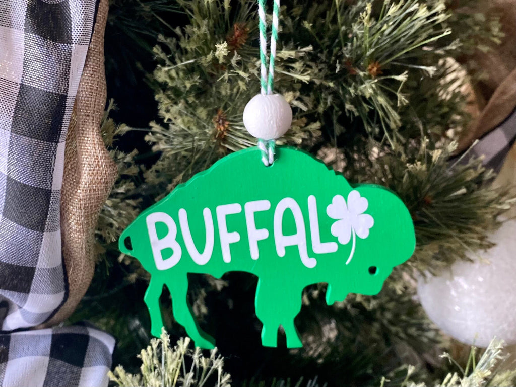 A green St. Patrick's Day buffalo ornament hanging from a Christmas tree, featuring the word 'BUFFAL' in white with a shamrock and a white bead at the top.