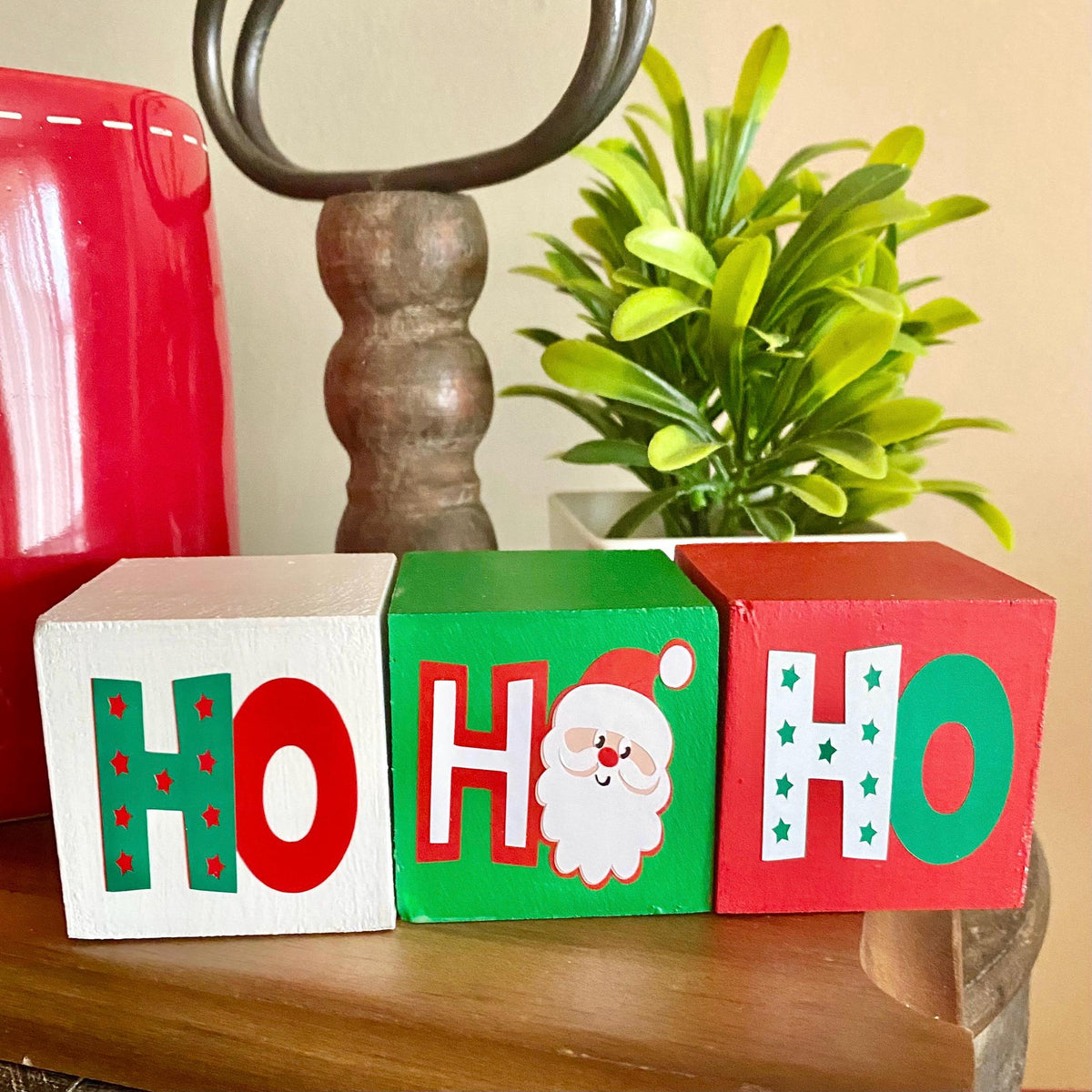 Three wooden HO blocks with Christmas-themed lettering and a Santa face, displayed on a wooden table next to a red mug and a potted plant.