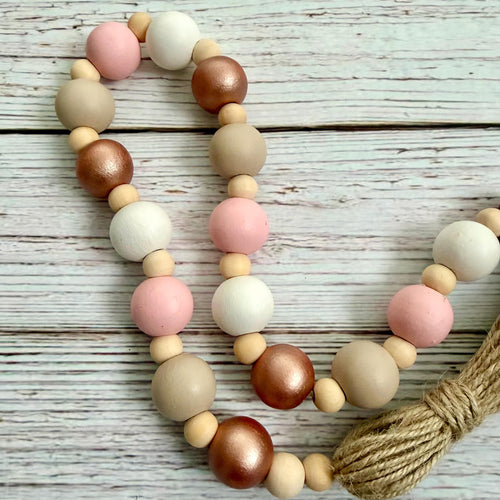 A beaded garland with alternating matte beige, white, and pink wooden beads, metallic rose gold and copper beads, and a jute rope end, displayed on a whitewashed wood background.