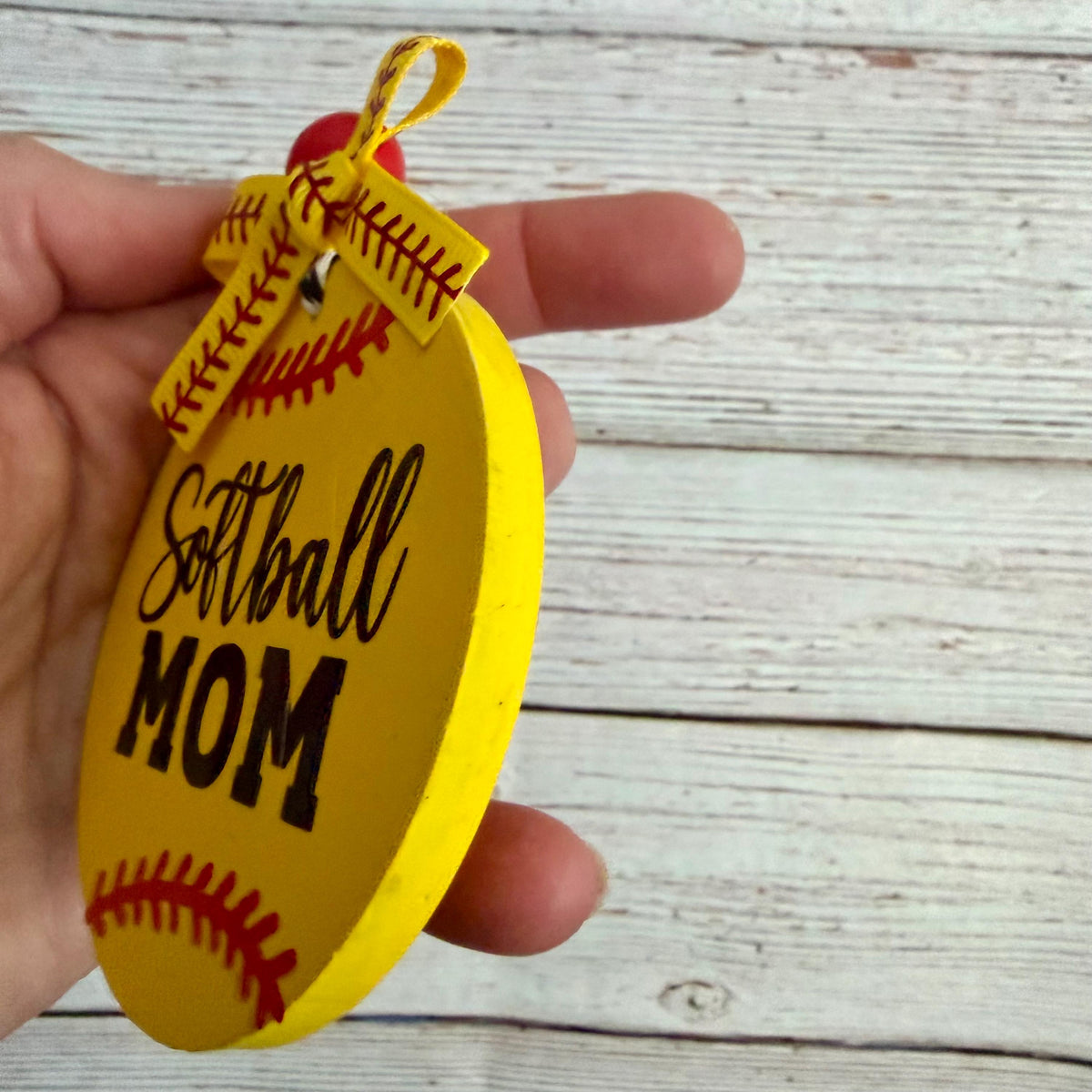 A hand holding a yellow oval wooden ornament shaped like a softball, printed with 'Softball MOM' in black script and block letters, with red baseball-style stitching details and a yellow hanging loop.