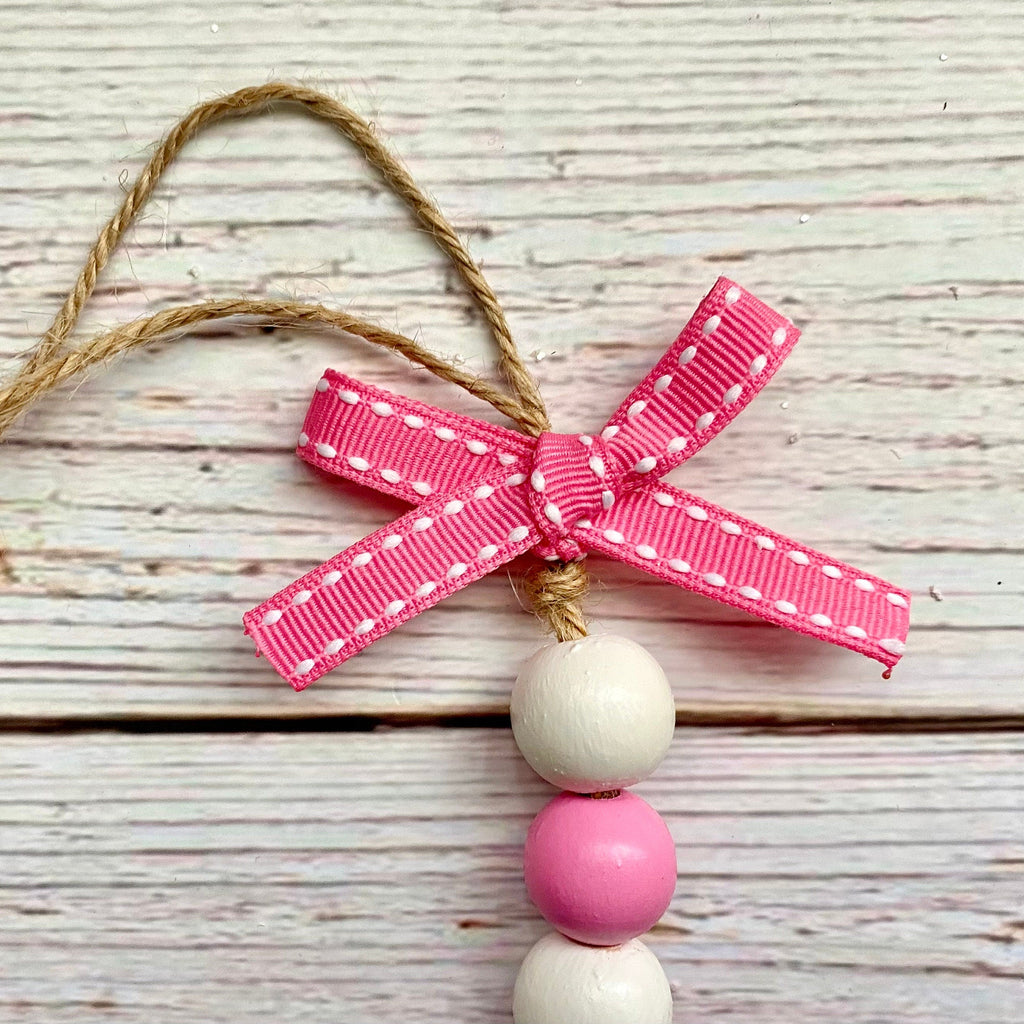 A decorative hanging ornament with a pink ribbon bow, white and pink wooden beads, and a tan string on a whitewashed wood plank background.