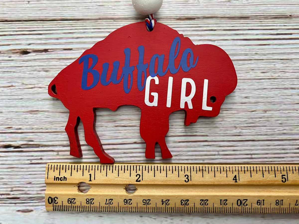 A red buffalo silhouette hanging ornament with the words 'Buffalo GIRL' in blue and white lettering, shown above a wooden ruler for scale on a whitewashed wood background.