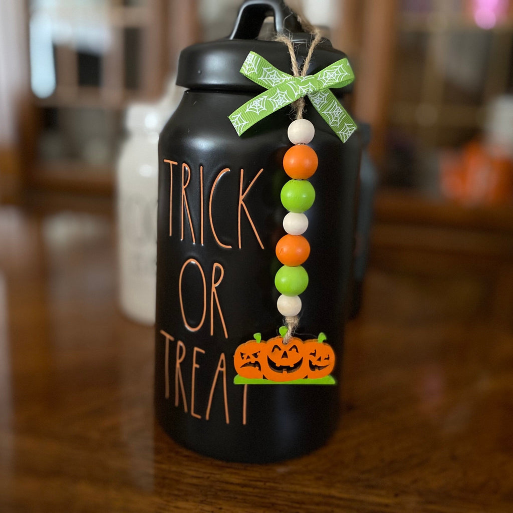 A black insulated water bottle with 'TRICK OR TREAT' written on the front in orange lettering, decorated with a green patterned ribbon bow and a hanging beaded garland with orange, white, and green beads and jack-o'-lanterns at the bottom, set on a wooden table.