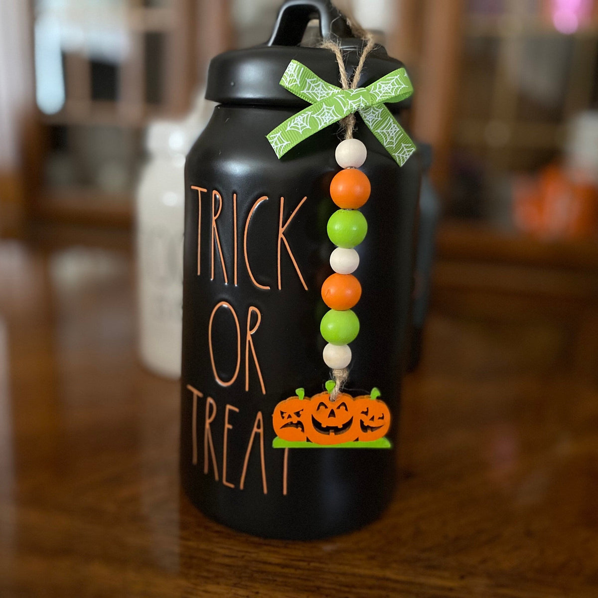 A black insulated water bottle with 'TRICK OR TREAT' written on the front in orange lettering, decorated with a green patterned ribbon bow and a hanging beaded garland with orange, white, and green beads and jack-o'-lanterns at the bottom, set on a wooden table.