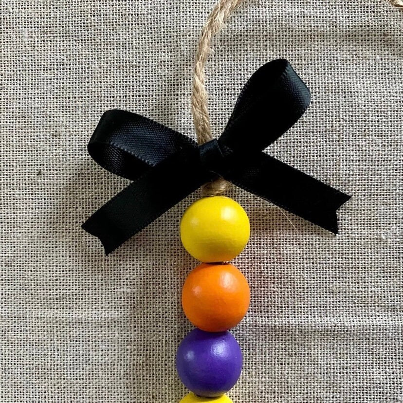 A handmade wooden bead ornament with a black ribbon bow and natural jute hanger, featuring yellow, orange, purple, and yellow wooden beads on a textured beige fabric background.