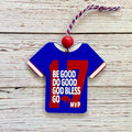 A blue t-shirt-shaped Christmas ornament with red and white trim, featuring the number 17 and the text 'BE GOOD DO GOOD GOD BLESS GOD BLESS GO MVP' with a small red buffalo graphic. It hangs from a red bead and red-and-white string against a whitewashed wood background.