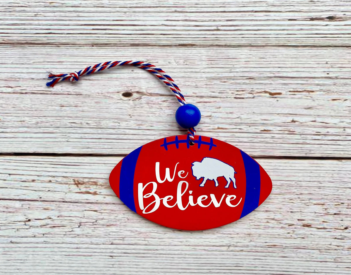 A red and blue wooden football-shaped ornament with white 'We Believe' text and a white bison graphic, hanging on a rustic white wood plank background.