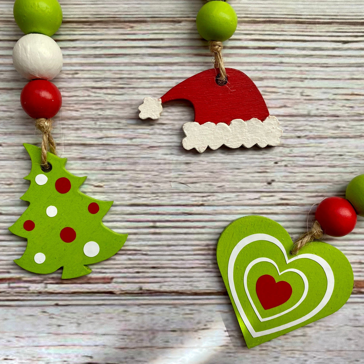 Three festive wooden bead garlands laid out on a whitewashed wood surface. One garland features a green Christmas tree cutout with red and white polka dots, another has a red Santa hat cutout, and the third shows a green heart cutout with white and red concentric hearts.
