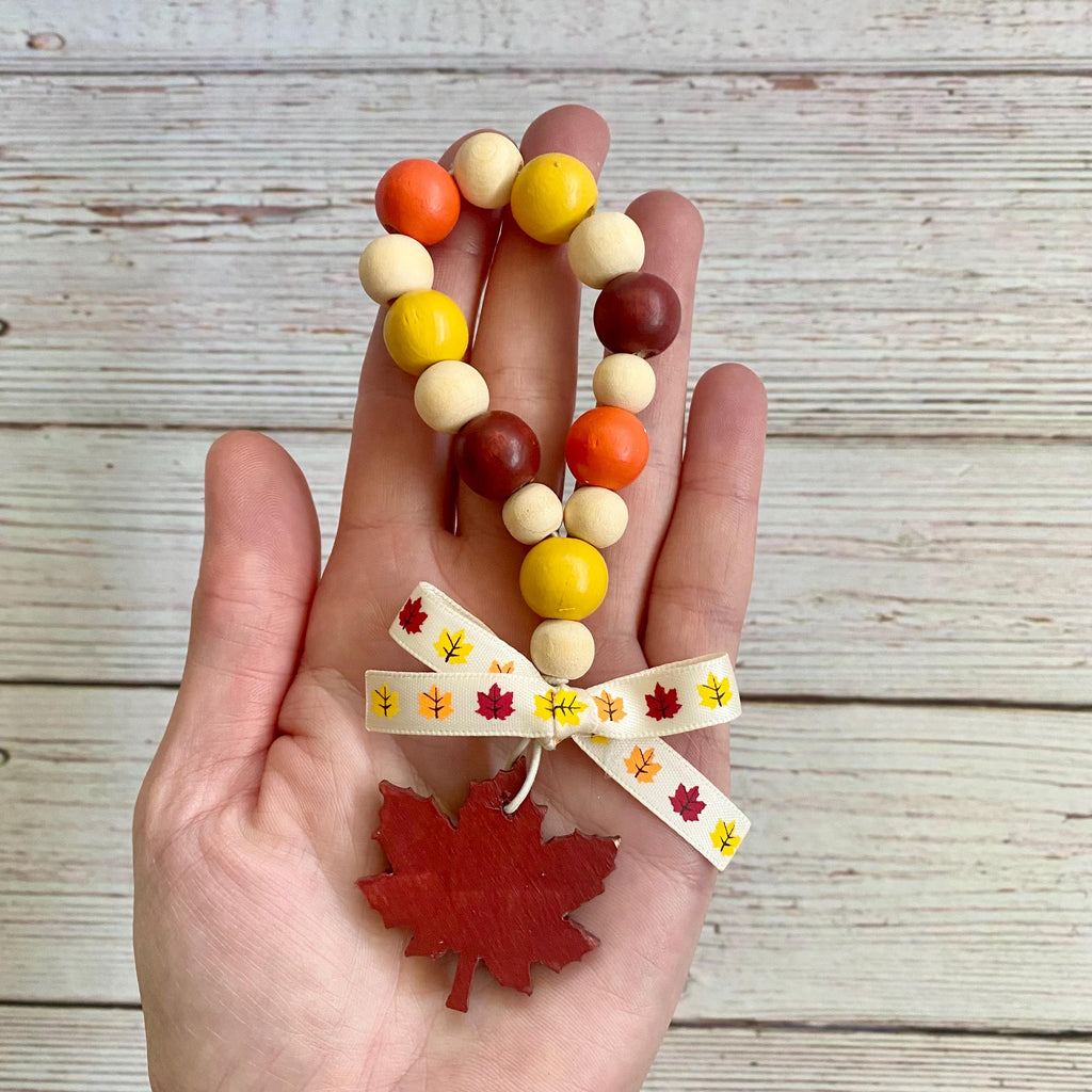 A hand holds a beaded loop with orange, yellow, cream, and brown round beads, tied with a cream ribbon printed with small autumn leaves, and finished with a red maple leaf charm.