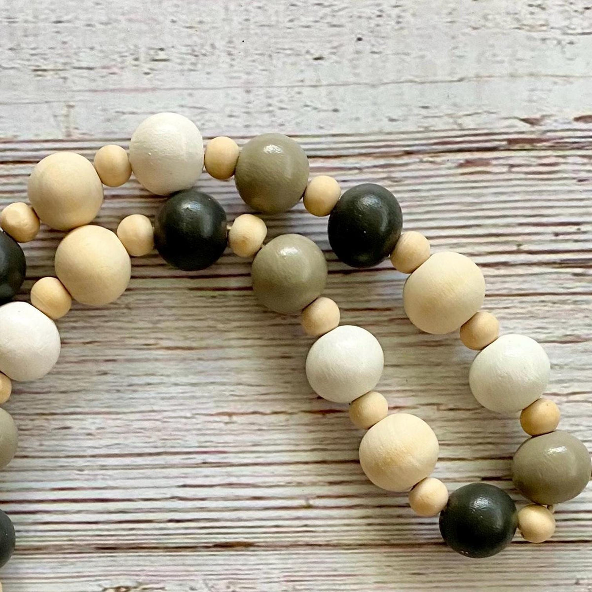 A close-up of a beaded strand with alternating matte wood beads in natural beige, olive green, and dark charcoal black, arranged on a whitewashed wooden surface.