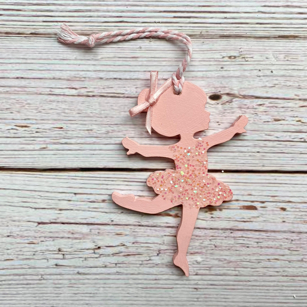 A pink ballerina-shaped ornament with a glittery dress and a pink-and-white string, displayed on a rustic, light gray wooden plank background.