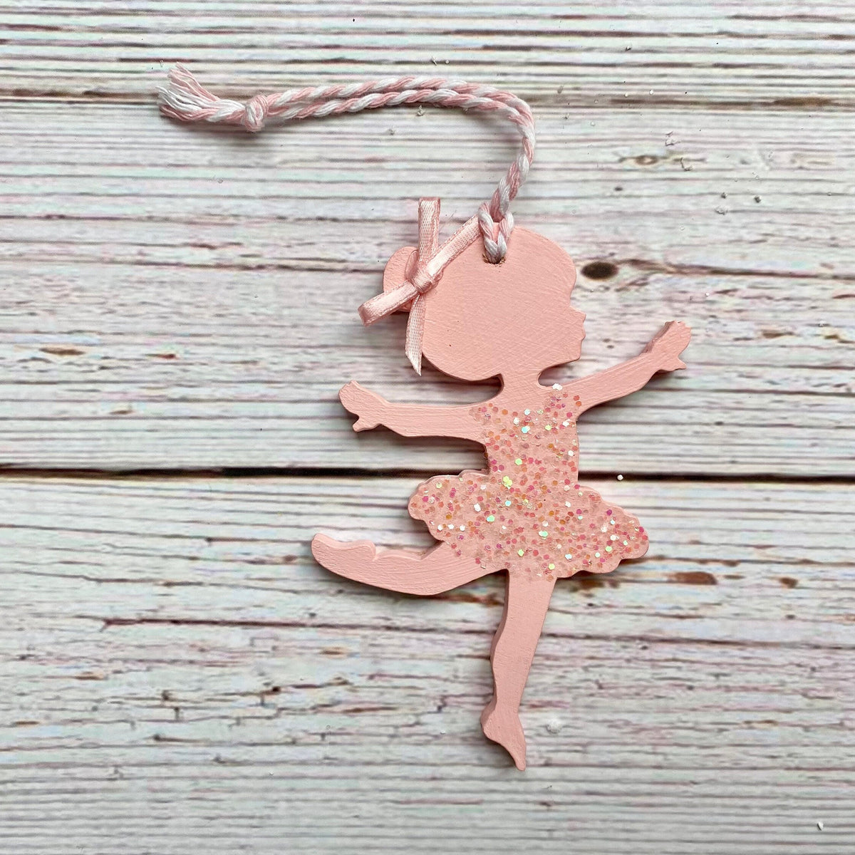 A pink ballerina-shaped ornament with a glittery dress and a pink-and-white string, displayed on a rustic, light gray wooden plank background.