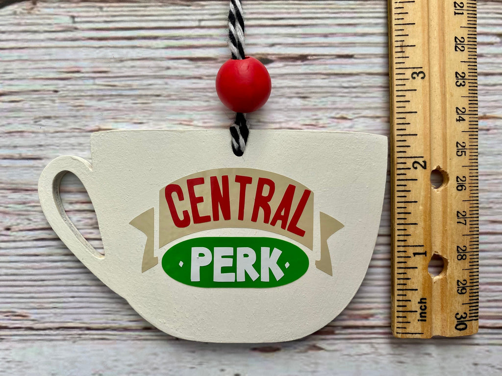 A hand-painted wooden ornament shaped like a teacup with a 'Central Perk' logo, shown next to a wooden ruler on a whitewashed wood background.