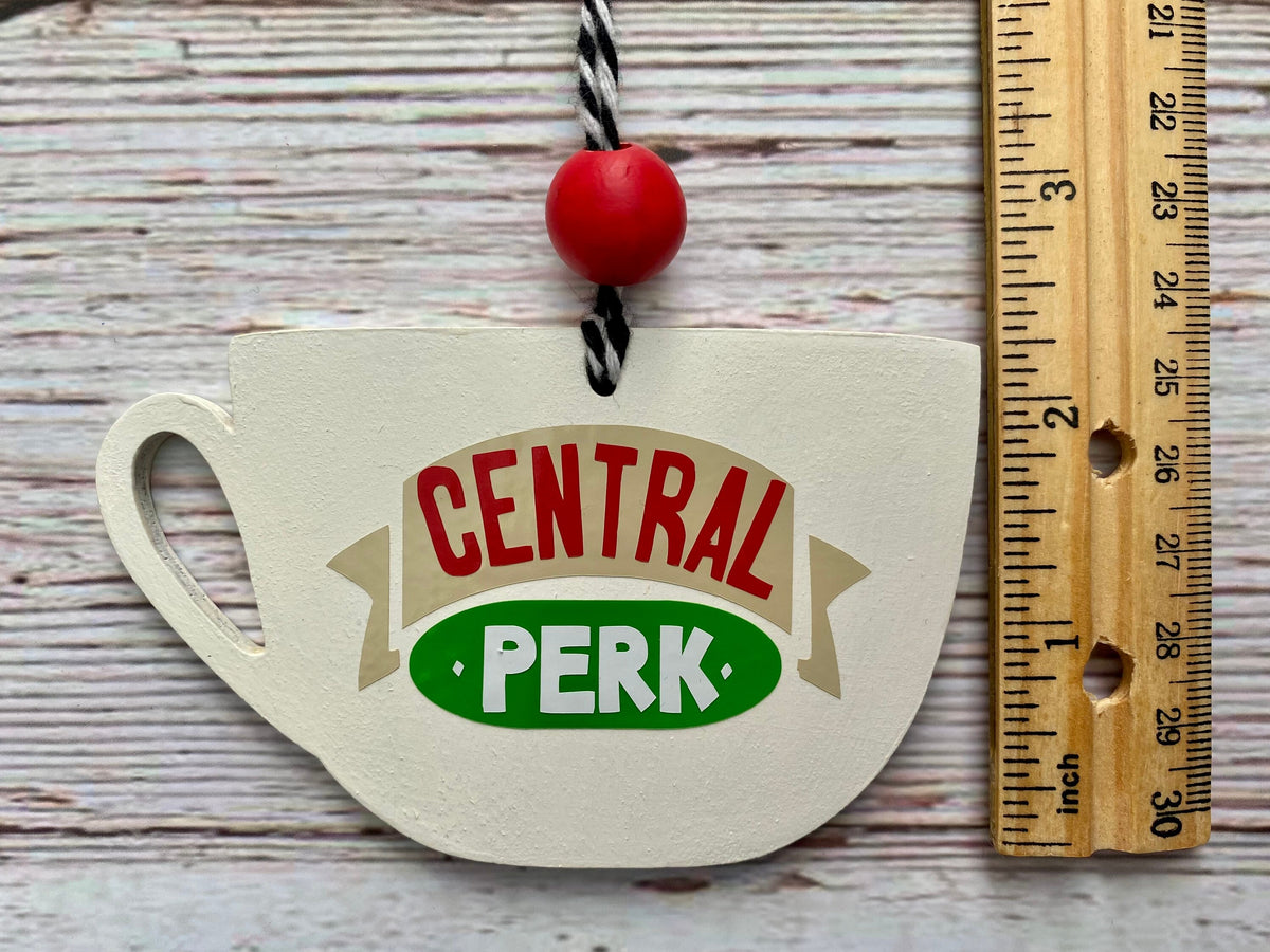 A hand-painted wooden ornament shaped like a teacup with a 'Central Perk' logo, shown next to a wooden ruler on a whitewashed wood background.