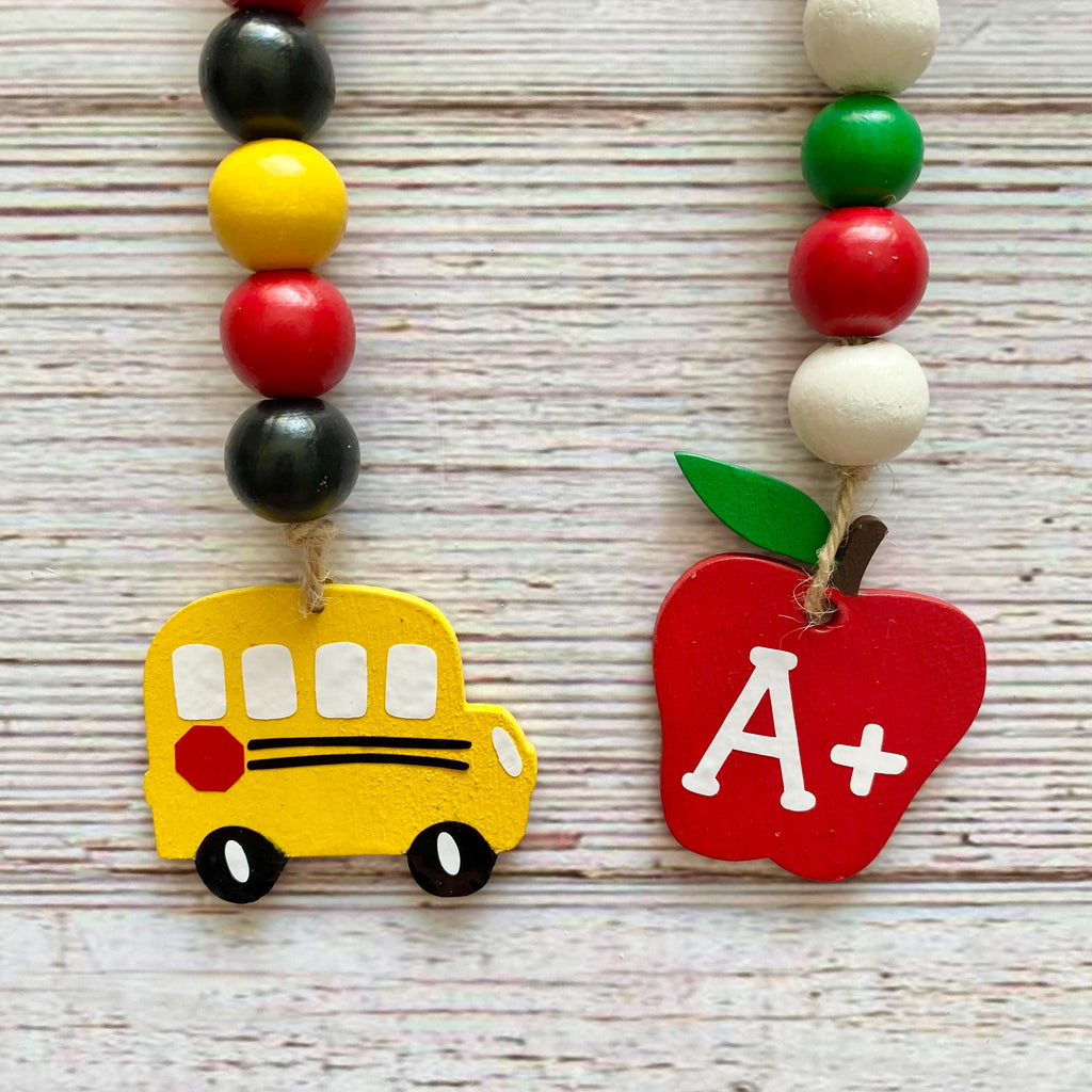 Two colorful beaded school-themed garlands displayed on a whitewashed wooden surface. One necklace features a yellow school bus charm with black and white details, and the other features a red apple charm with a white 'A+' lettering and a green leaf.