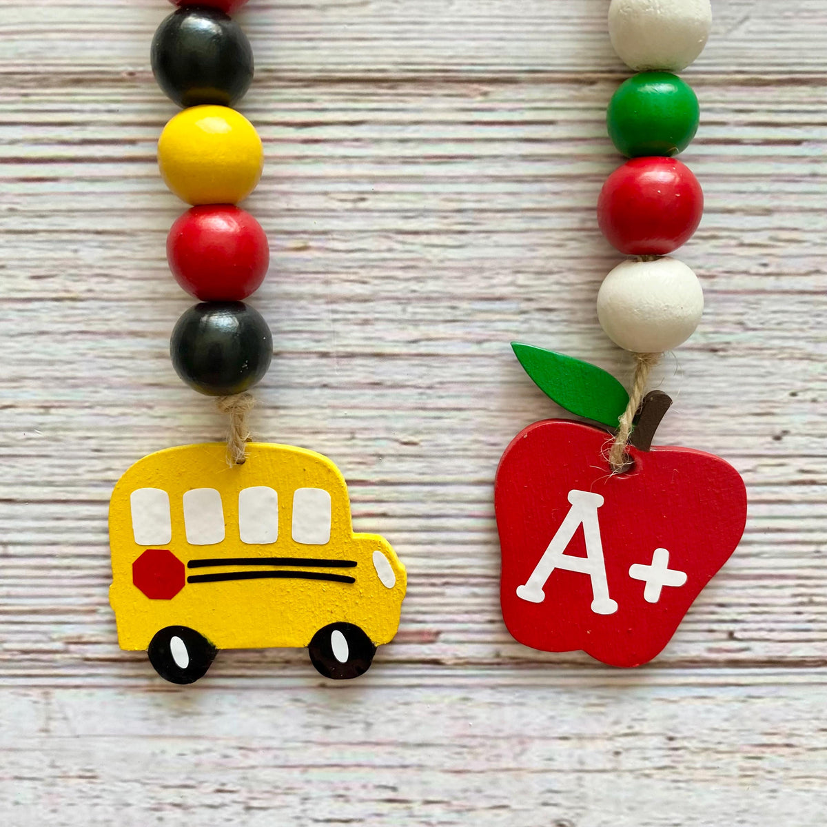 Two colorful beaded school-themed garlands displayed on a whitewashed wooden surface. One necklace features a yellow school bus charm with black and white details, and the other features a red apple charm with a white 'A+' lettering and a green leaf.