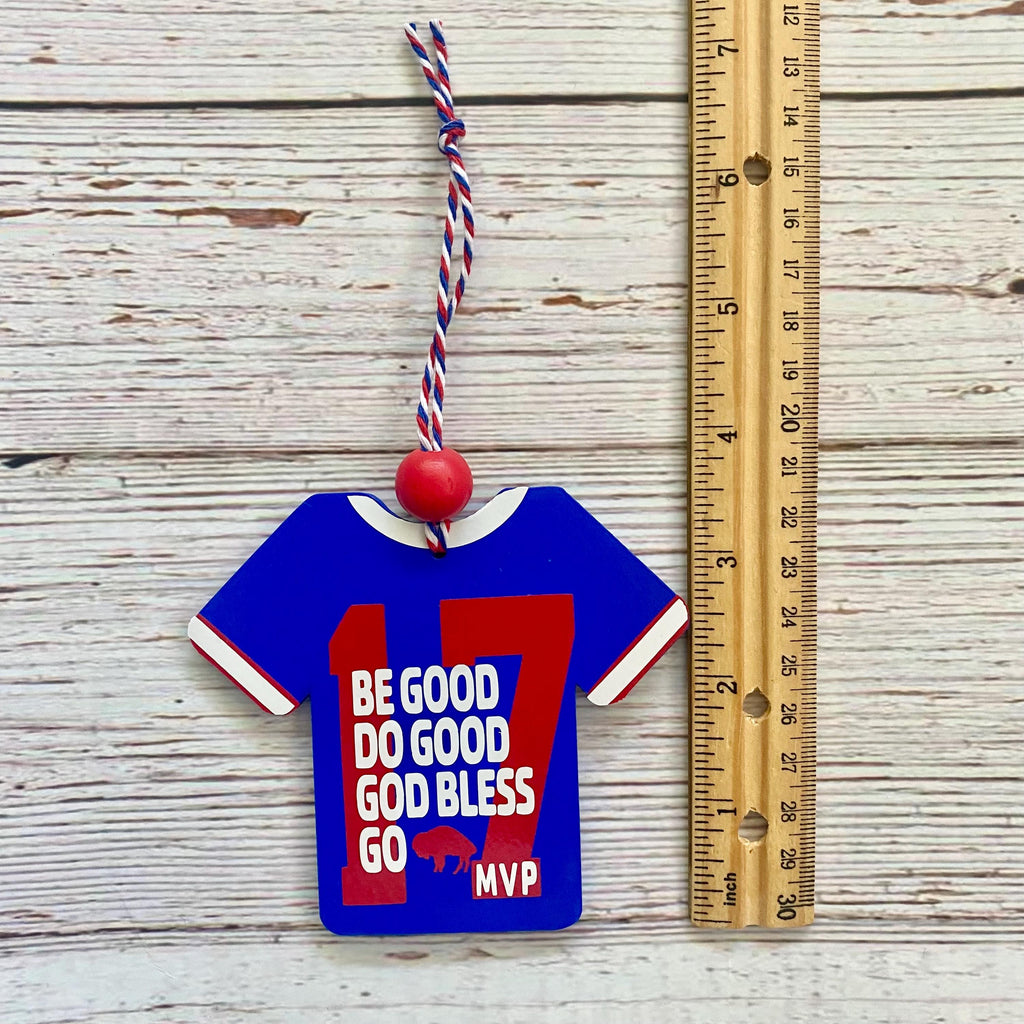 A blue and red jockey-shirt-shaped ornament with white text reading 'BE GOOD DO GOOD GOD BLESS GO MVP' and a red '17', hanging by a red, white, and blue string next to a wooden ruler on a whitewashed wood background.