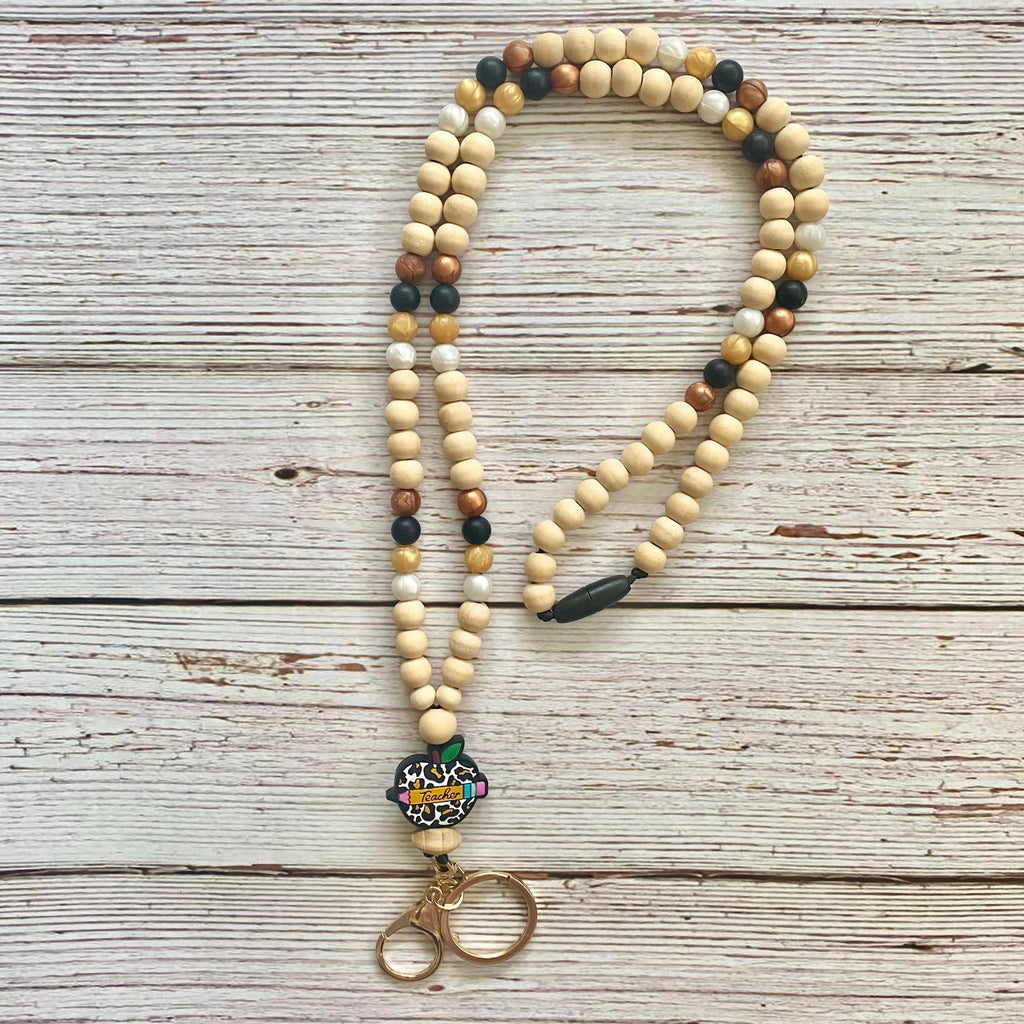 A beaded necklace with natural wood beads in cream, tan, and brown, accented with small black and dark brown beads, a colorful patterned charm, and a gold-tone metal clasp, displayed on a rustic white-washed wooden background.