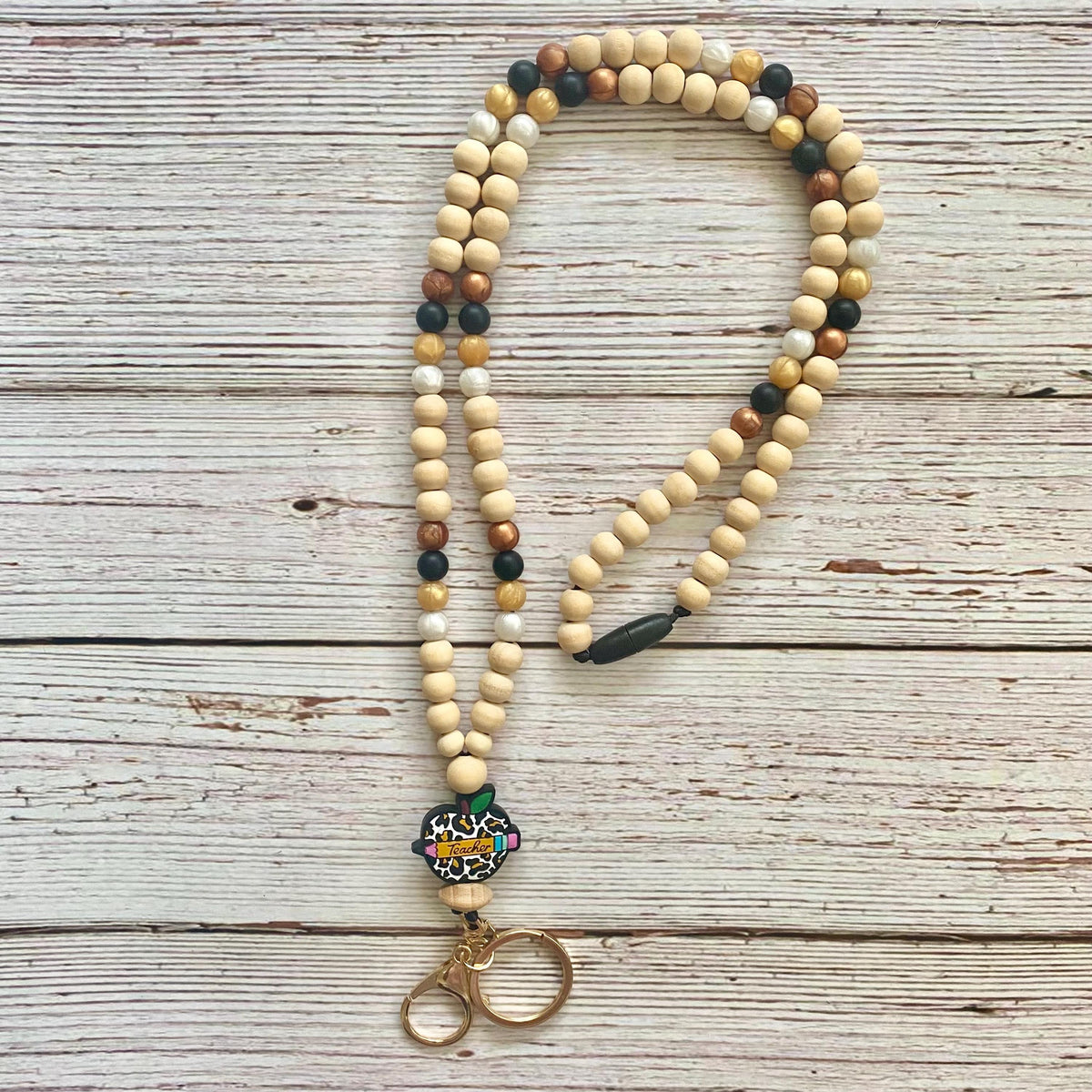 A beaded necklace with natural wood beads in cream, tan, and brown, accented with small black and dark brown beads, a colorful patterned charm, and a gold-tone metal clasp, displayed on a rustic white-washed wooden background.