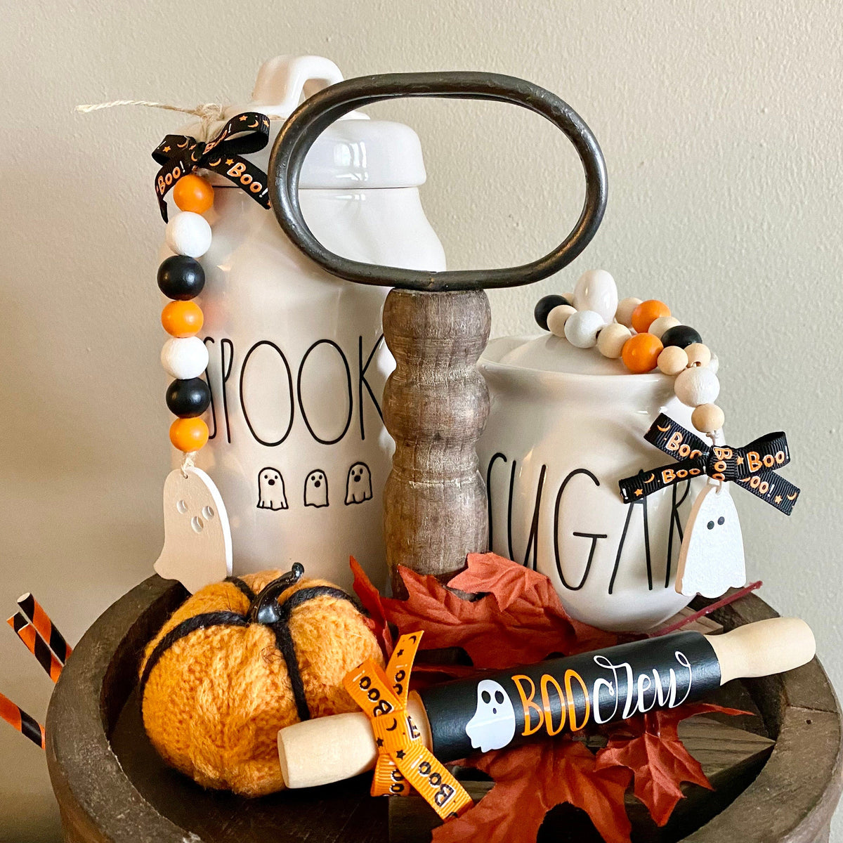 A styled Halloween-themed tabletop arrangement featuring two white ceramic canisters with 'BROOK' and 'SUGAR' lettering, decorated with black ribbon bows, ghost beads, a magnifying glass, a small orange pumpkin, fall leaves, and a rolling pin on a wooden surface against a plain beige wall.