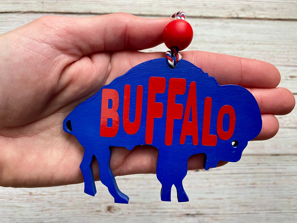 A hand holding a blue buffalo-shaped ornament with the word 'BUFFALO' in red letters, featuring a red bead at the top, set against a light wood background.