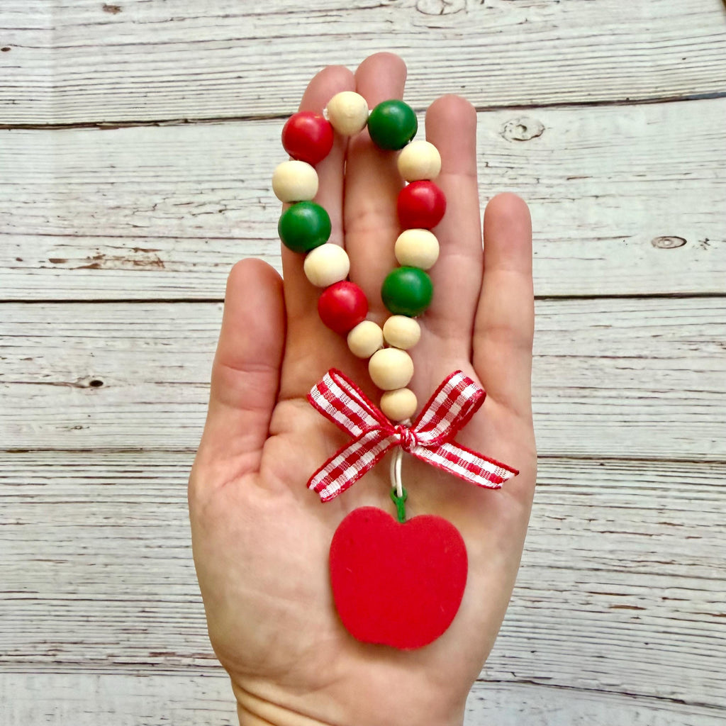 A hand holds a beaded loop  with red, green, and cream wooden beads, a red-and-white gingham ribbon bow, and a red apple-shaped hanging ornament, set against a rustic white wood plank background.