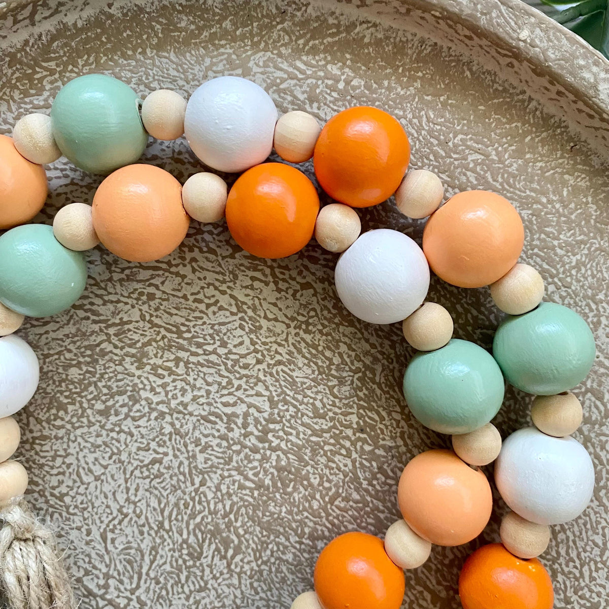 A close-up of a beaded strand featuring alternating large pastel beads in mint green, peach, orange, and white, interspersed with smaller natural wood beads, arranged on a textured beige tray