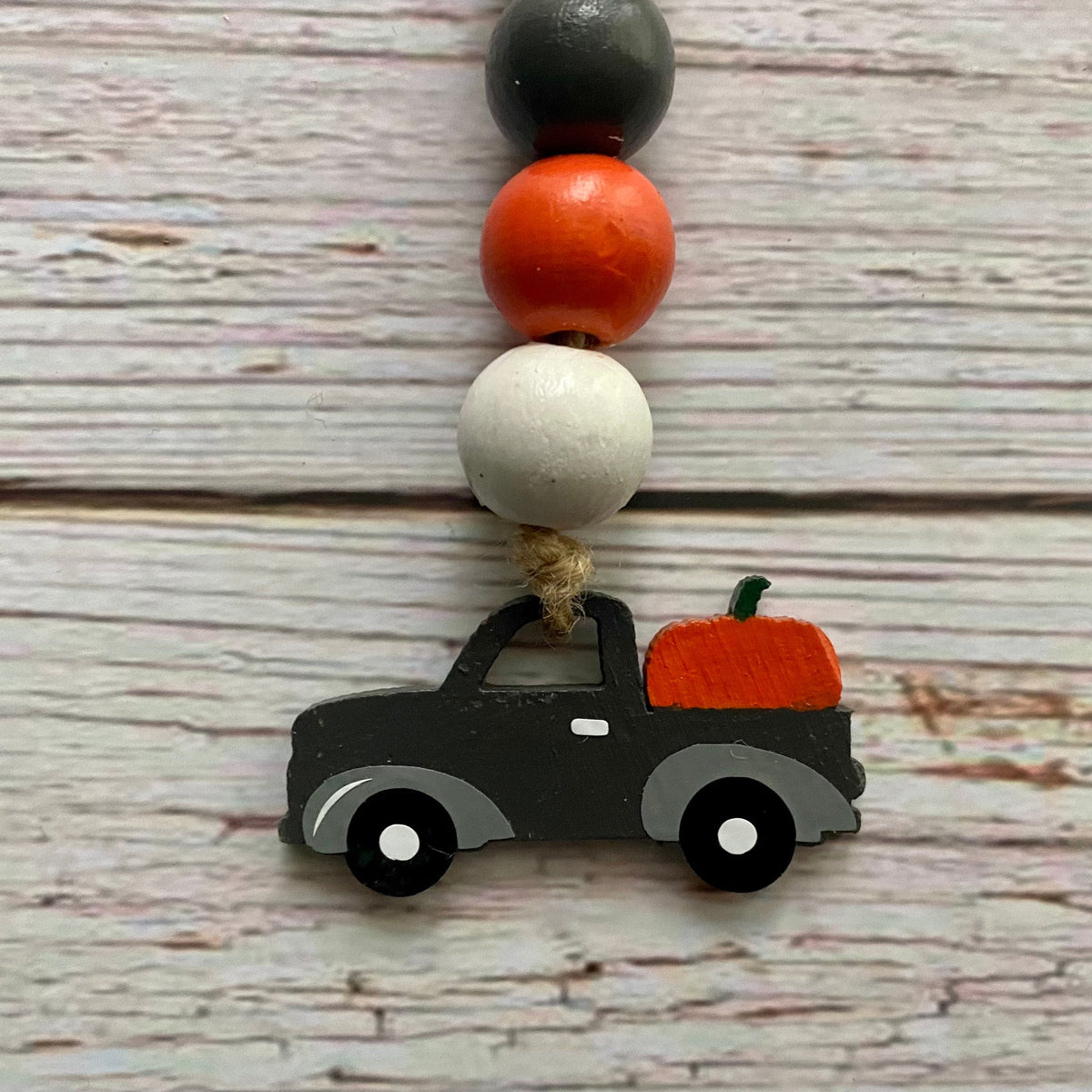 A beaded mini garland featuring a black wooden truck carrying an orange pumpkin, suspended from a string with four decorative wooden beads in black, orange, white, and gray, set against a rustic whitewashed wood background.