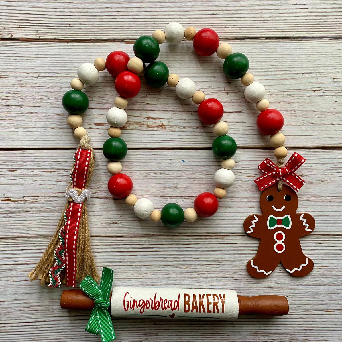 A Christmas-themed beaded garland with red, green, and white wooden beads, a gingerbread man ornament, a tassel, and a wooden rolling pin charm that reads 'Gingerbread BAKERY', displayed on a rustic white wood background.