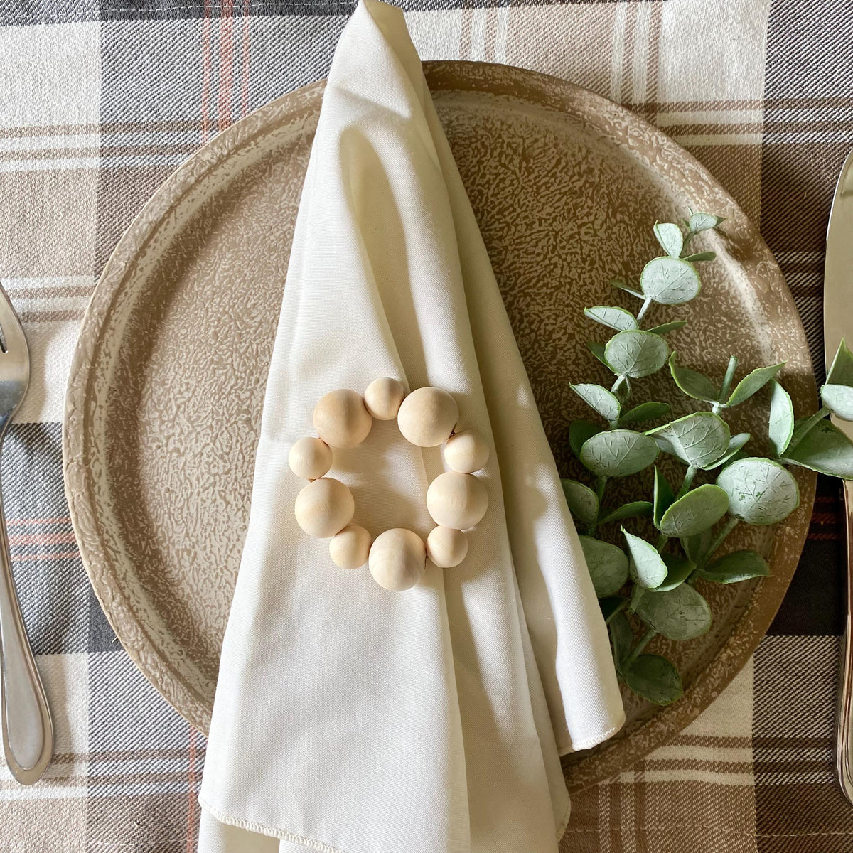 A styled tabletop setting with a beige ceramic plate, a cream folded napkin, a ring of natural wood beads, and green eucalyptus leaves on a plaid tablecloth.
