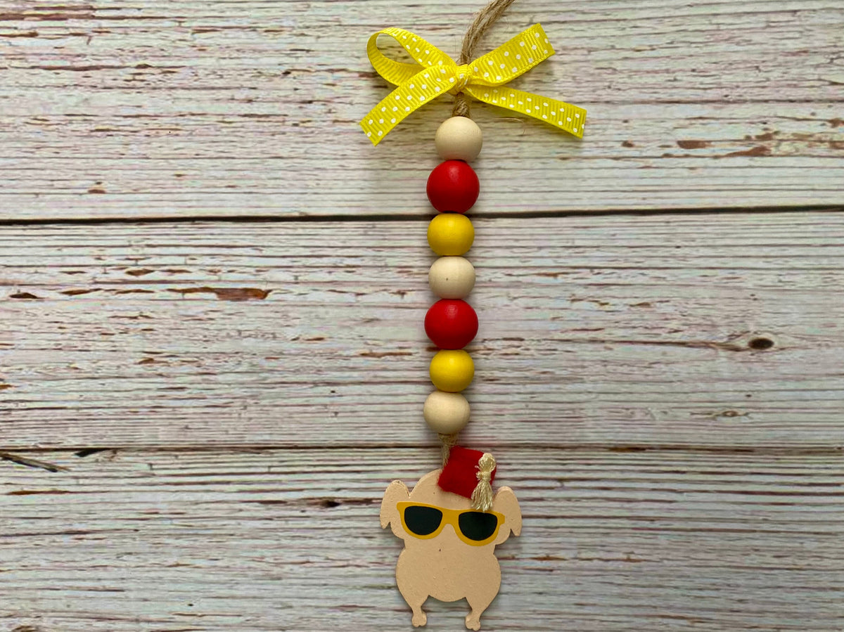 A handmade wooden bead hanging ornament featuring red and yellow wooden beads, a yellow polka-dot ribbon bow at the top, and a small wooden turkey charm wearing black sunglasses and a red Santa hat, displayed against a rustic white-washed wood background.
