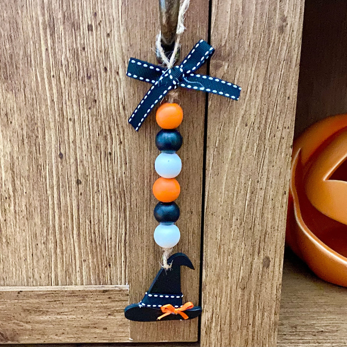 A handmade wooden bead hanging ornament with a navy blue ribbon bow, orange and light blue beads, and a small painted black witch hat, displayed on a rustic wooden shelf next to a jack-o'-lantern.