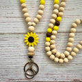 A wooden bead keychain or lanyard-style accessory laid on a whitewashed wood surface. The design features natural light wood beads, bright yellow accent beads, a yellow sunflower charm with a dark center, and a metal key ring at the bottom.