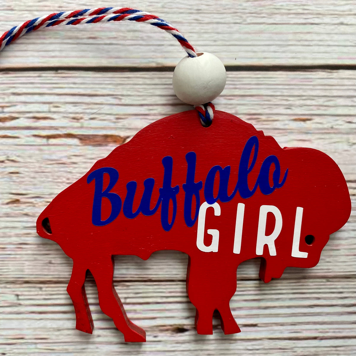 A red wooden ornament shaped like a buffalo silhouette with the words 'Buffalo GIRL' in blue and white lettering, hanging from a red, white, and blue string with a white bead, set against a whitewashed wood background.