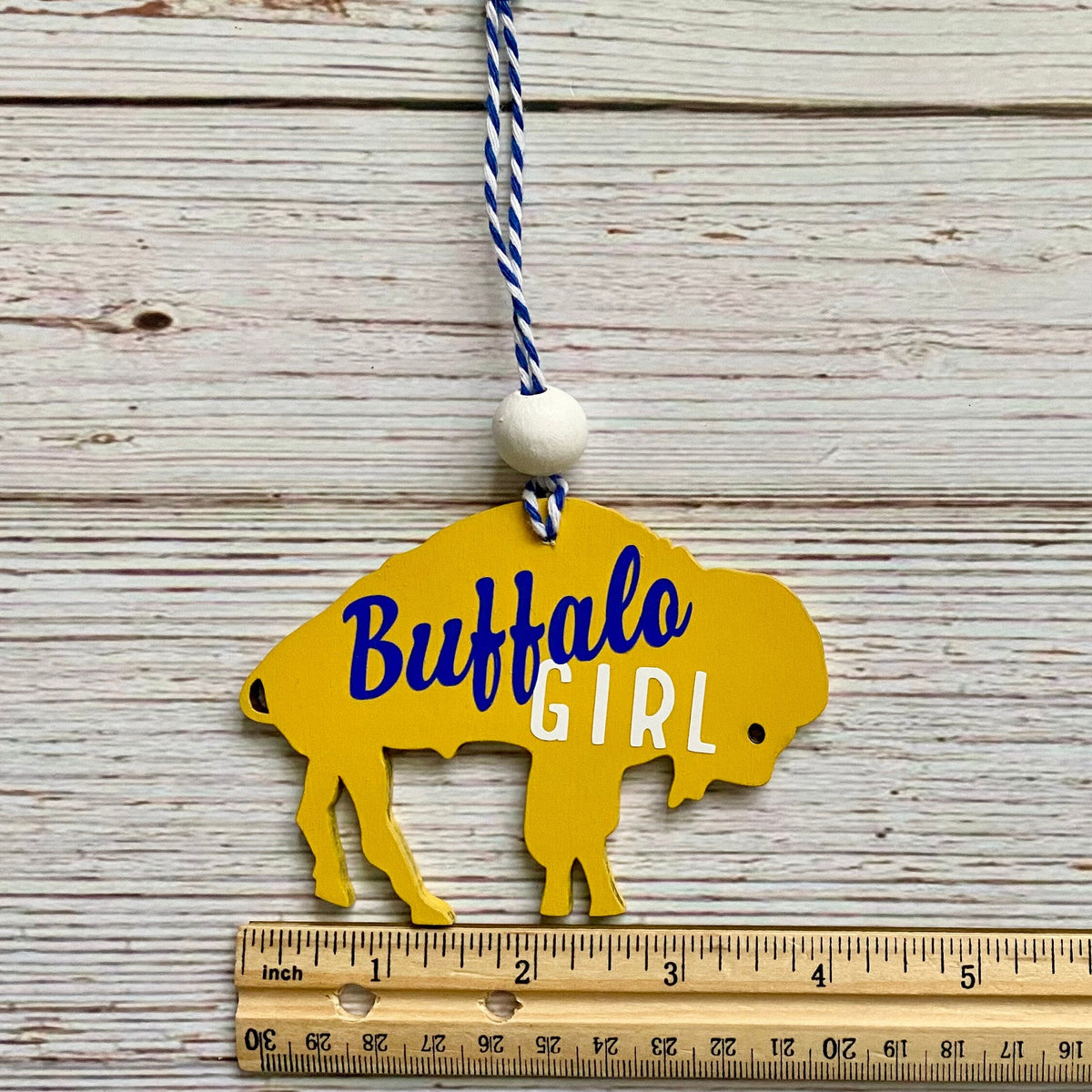 Yellow wooden Buffalo silhouette ornament with 'Buffalo GIRL' text, hanging from a blue-and-white string on a whitewashed wood background with a ruler below for scale.