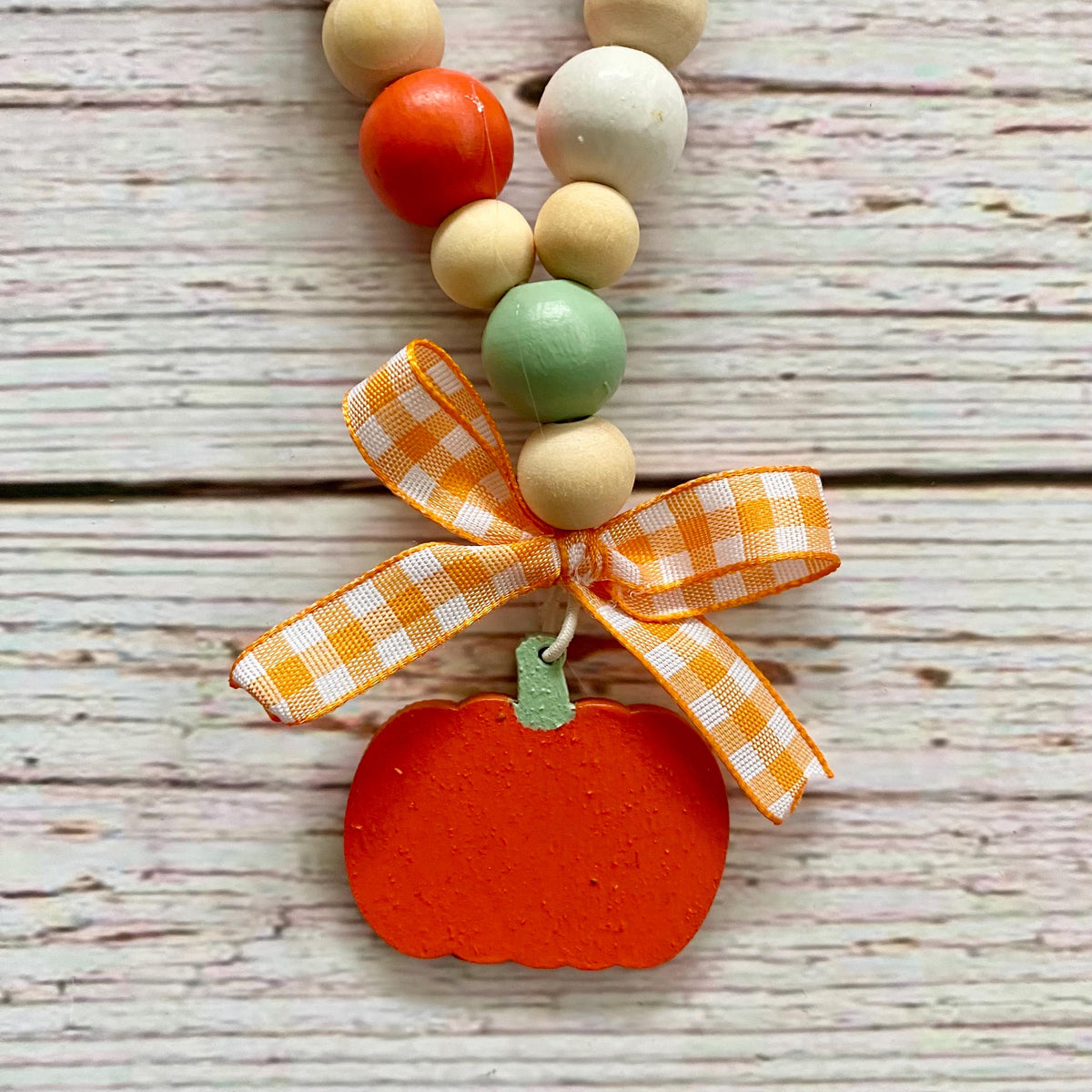 A wooden bead garland featuring a hand-painted orange pumpkin charm, tied with an orange and white gingham ribbon bow, displayed on a rustic white-washed wooden background.