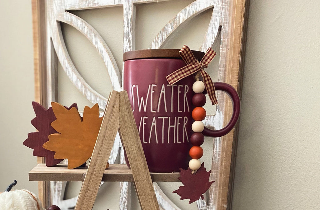 A burgundy enamel mug with a wooden lid and white text reading 'SWEATER WEATHER,' styled on a small wooden stand with fall leaves and a bead garland against a rustic white window frame.